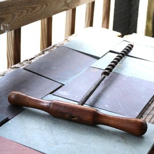 May include: A wooden rolling pin with a dark finish, lying on a slate tile surface. A metal corkscrew-style tool is also present. The items are likely kitchen or bar tools.