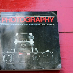 Puede incluir: Un libro en blanco y negro titulado "Fotografía" de Barbara London Upton con John Upton, tercera edición. La portada presenta un primer plano de un objetivo de cámara.