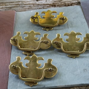 Op de afbeelding: Een verzameling van vijf antieke messing lade handgrepen met een sierlijk ontwerp. De handgrepen zijn gerangschikt op een grijze ondergrond, wat hun verouderde, gouden afwerking laat zien. Deze vintage hardware stukken zijn ideaal voor meubelrestauratie.