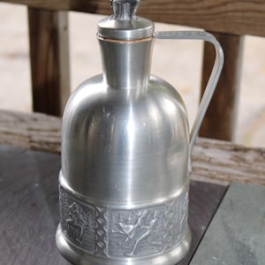 May include: A silver pewter pitcher with a detailed design of figures and a lid featuring a figurine of a person playing a musical instrument.