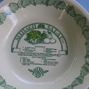 Puede incluir: Un bol de ensalada de cerámica blanca con un borde verde y una receta de ensalada de espinacas impresa en el interior. La receta incluye ingredientes como espinacas, tocino, huevos, champiñones y un aderezo ácido.