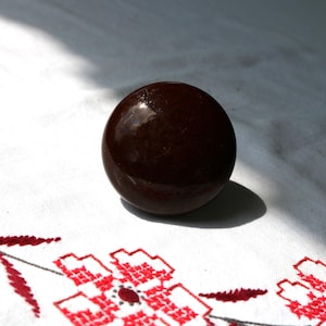 May include: A dark brown, spherical object with a glossy finish. The object sits on a white cloth with red floral embroidery. The lighting creates a shadow to the right of the sphere.