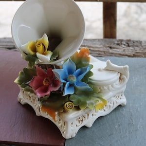 May include: A white porcelain figurine of a vintage gramophone with a floral design. The gramophone is decorated with yellow, red, blue, and green flowers. The base of the gramophone is ornately decorated.