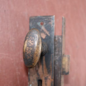 Puede incluir: Primer plano de un pomo y pestillo de puerta antiguos. El pomo de forma ovalada tiene un acabado bronce desgastado. La placa metálica y el pestillo muestran signos de edad y desgaste, con una pátina oscura y envejecida. El fondo es rojo apagado.