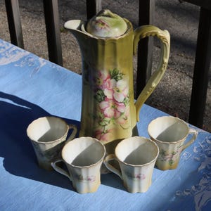 Puede incluir: Juego de té vintage con una cafetera verde oliva con tapa y cuatro tazas a juego. El juego está adornado con diseños florales rosas y blancos. Las tazas y la cafetera tienen un borde festoneado.