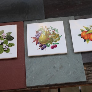 May include: Three square ceramic tiles with fruit and floral designs. One tile features pears, grapes, and cherries. Another shows oranges and blossoms. The third tile displays white flowers and green leaves. The tiles are on a dark surface.