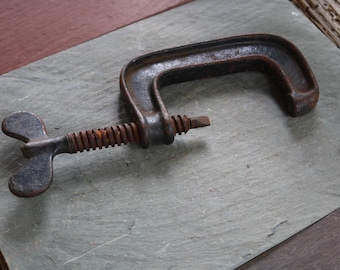 May include: An antique C-clamp, made of dark metal, rests on a gray stone surface. The clamp features a threaded screw mechanism with a butterfly-shaped handle for tightening. The metal shows signs of age and rust.