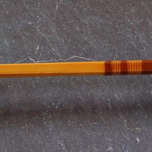 K&ouml;nnte beinhalten: Eine Angelrute aus Holz mit braunen und orangen Streifen. Die Rute ist aus Bambus und hat einen Metallrollenhalter.