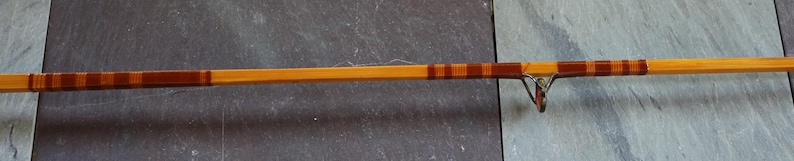 K&ouml;nnte beinhalten: Eine Angelrute aus Holz mit braunen und orangen Streifen. Die Rute ist aus Bambus und hat einen Metallrollenhalter.