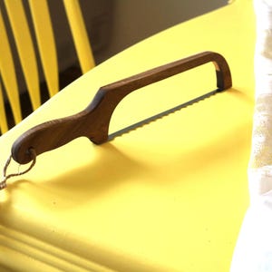 Puede incluir: Un cuchillo de pan de madera con un mango curvo y una hoja dentada descansa sobre una superficie amarilla. El cuchillo es de madera oscura y tiene un acabado natural. Un trozo de cuerda está unido al mango. El fondo incluye una silla amarilla y tela blanca.