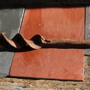 Puede incluir: Una broca de metal oxidada con un diseño en espiral, que se encuentra sobre una superficie de madera de color gris oscuro, rojo y marrón.