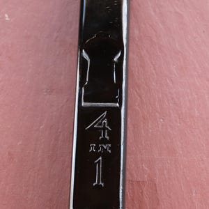 May include: A black, rectangular bottle with a keyhole-shaped indentation and the numbers "4, RM, 1" embossed on the front. The bottle has a glossy finish and is set against a reddish-brown background.