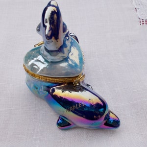 May include: A blue and iridescent porcelain trinket box shaped like a dolphin with a heart-shaped lid. The lid features a smaller dolphin figurine. The dolphin's body is inscribed with "Naples, FL."