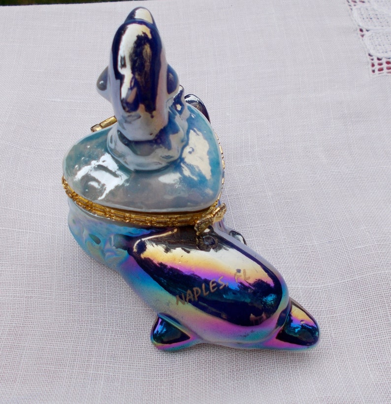 May include: A blue and iridescent porcelain trinket box shaped like a dolphin with a heart-shaped lid. The lid features a smaller dolphin figurine. The dolphin's body is inscribed with "Naples, FL."