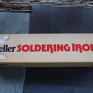Puede incluir: Caja rectangular para un soldador Weller. La caja es de color beige con letras rojas que dicen "Weller SOLDERING IRON". La caja está sobre una superficie gris oscuro.