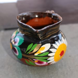 May include: A small, hand-painted ceramic pitcher with a black exterior. The pitcher is adorned with colorful floral designs in white, yellow, green, and purple. The rim features a dark brown and gold striped pattern. A charming piece for kitchen decor.