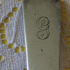 Può includere: Primo piano di un'utensile d'argento, forse un cucchiaio o una forchetta, con un'estremità arrotondata. L'utensile ha una lettera "G" decorativa e corsiva incisa sulla superficie. Lo sfondo è un tessuto strutturato bianco sporco.