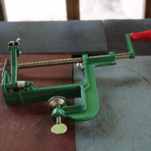May include: A green metal apple peeler and corer featuring a horizontal threaded rod, a sharp three-pronged fork, a silver-toned screw clamp base for mounting, and a red plastic handle attached to a crank for manual operation.