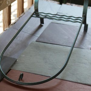 May include: A dark green metal stand featuring a U-shaped frame with a small hook at the front, four vertical legs, and a horizontal support bar with a wavy, undulating design connecting the two rear legs for structural stability.