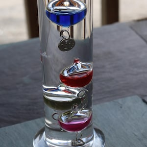 May include: A clear glass Galileo thermometer with four colored glass bulbs filled with liquid. The bulbs are blue, red, green, and pink. The thermometer sits on a clear base and has a small metal tag attached to each bulb.