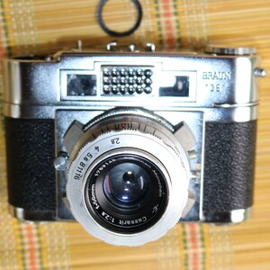 Pu&ograve; includere: Una macchina fotografica a pellicola da 35 mm argentata e nera con la scritta "BRAUNY 35" sulla parte superiore. La macchina fotografica ha un obiettivo argentato con il testo "COSSINA 1:2.8 50mm" e "VETERAN" stampato su di esso.