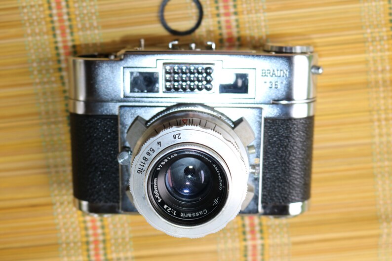 Pu&ograve; includere: Una macchina fotografica a pellicola da 35 mm argentata e nera con la scritta "BRAUNY 35" sulla parte superiore. La macchina fotografica ha un obiettivo argentato con il testo "COSSINA 1:2.8 50mm" e "VETERAN" stampato su di esso.