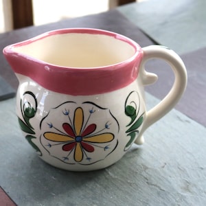 May include: A small, white ceramic pitcher with a pink rim and handle. The pitcher is decorated with a floral design in yellow, red, green, and blue. The pitcher has a spout for easy pouring.