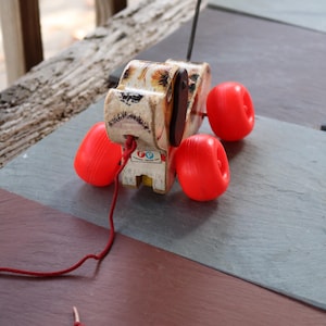 Op de afbeelding: Een vintage houten trekdier in de vorm van een hond met rode wielen en een touwtje eraan vast. De hond heeft een bruine leren neus en zwarte ogen. Het speelgoed ligt op een leisteen oppervlak.