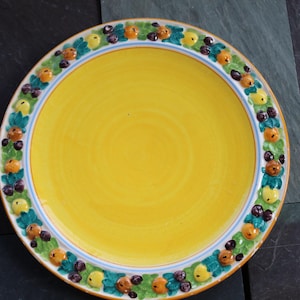 May include: A yellow ceramic plate with a decorative border of green leaves, orange and purple fruit, and a white rim.