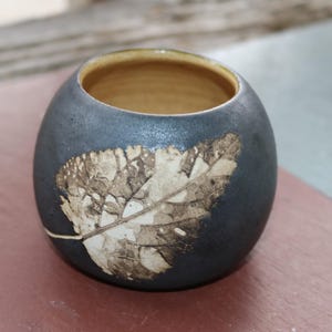 Può includere: Un piccolo vaso rotondo in ceramica nera con un disegno a foglia bianca. Il vaso ha una finitura liscia e opaca e un bordo leggermente arrotondato.