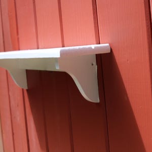May include: A white wooden wall shelf featuring a flat rectangular top surface supported by two curved brackets underneath. The shelf is mounted against a vertical red wooden plank wall with visible grain and vertical seams between the boards.