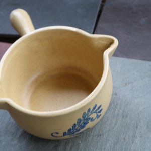 May include: A beige ceramic sauce bowl with a handle and a pouring spout. The bowl has a blue floral design on the side. The bowl is sitting on a gray surface.