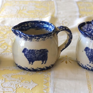 May include: A cream-colored ceramic creamer, sugar bowl with lid, and a small jar with a lid. Each piece features a blue sheep silhouette and blue speckled trim. The set is displayed on a white and yellow patterned tablecloth.