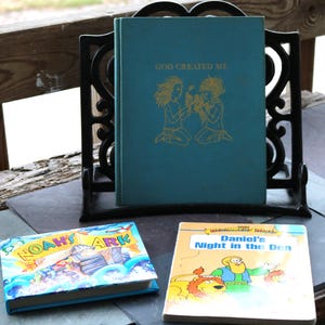 Może przedstawiać: Trzy książki dla dzieci na drewnianym stole. Górna książka jest niebieska i ma tytuł "God Created Me". Środkowa książka jest niebieska i ma tytuł "Noah's Ark". Dolna książka jest żółta i ma tytuł "Daniel's Night in the Den".