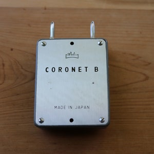 May include: A silver metal box with the text "CORONET B" and "MADE IN JAPAN" on the front. The box has two metal loops on the top.