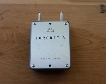 May include: A silver metal box with the text "CORONET B" and "MADE IN JAPAN" on the front. The box has two metal loops on the top.