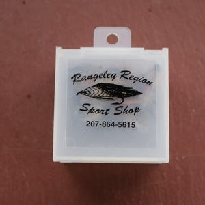May include: A white plastic box with a clear lid and a black and white logo that says "Rangeley Region Sport Shop" and a phone number "207-864-5615".