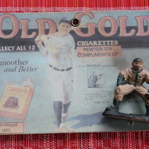 Könnte beinhalten: Vintage "Old Gold" Zigarettenwerbung mit Baseball-Motiv. Die Werbung zeigt eine Baseballspieler-Figur und den Text "Old Gold Cigarettes". Das Copyright-Datum ist 1953.