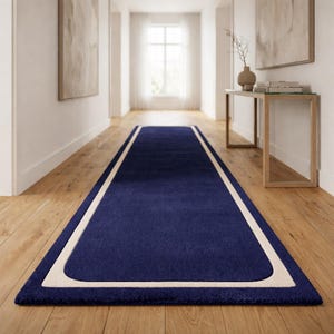 May include: A navy blue runner rug with a cream-colored border. The rectangular rug is placed on a hardwood floor in a hallway. The rug's dimensions are not visible in the image.