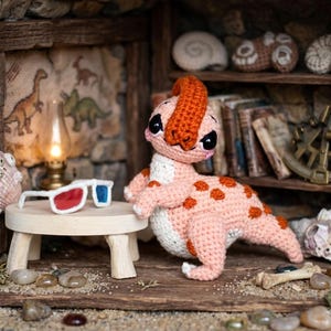 May include: A handmade crochet dinosaur in shades of pink and orange, with a white belly and orange spots. It is posed next to a small wooden table with 3D glasses. The scene is set in a miniature diorama with a dinosaur theme.