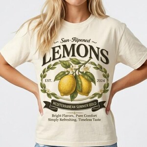 May include: Cream-colored t-shirt featuring a graphic of two yellow lemons on a branch with leaves and flowers. The word "LEMONS" is above the lemons, with "Sun-Ripened" above and "EST. 2024" below. The text "MEDITERRANEAN SUMMER GOLD" is on a banner.