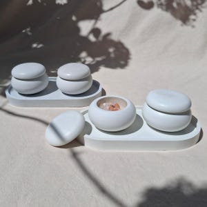 Könnte beinhalten: Zwei weiße ovale Tabletts mit jeweils zwei glatten, abgerundeten weißen Behältern mit Deckel. Ein Behälter ist geöffnet und zeigt kleine rosa Salzkristalle im Inneren. Die Behälter haben eine matte Oberfläche und eine kieselartige Form.