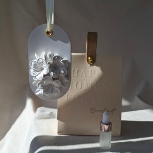 Könnte beinhalten: Ein weißes, ovales Hängeornament mit einem Relief aus weißen Blumen, aufgehängt an einem weißen Band. Daneben steht eine beige Geschenktüte mit dem geprägten Text HAPPY MOMENT FOR YOU Sweet gift und eine kleine Glastropfflasche.