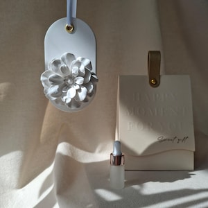 Könnte beinhalten: Ein weißes, ovales Hängeornament mit einer skulpturalen 3D-Blume, eine kleine Glastropfflasche und ein cremefarbenes, rechteckiges Geschenkanhänger mit Lederband. Der Anhänger ist mit HAPPY MOMENT FOR YOU und Sweet gift geprägt.