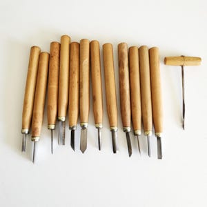 May include: A collection of wood carving tools with wooden handles and metal blades. The set includes various gouges and a corkscrew, all arranged on a white surface. The tools are designed for detailed woodworking and carving projects.