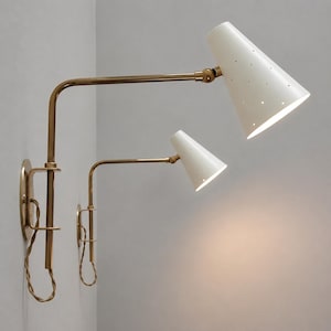 May include: Two wall-mounted lamps with a mid-century modern design. The lamps feature white, cone-shaped shades with small perforations and brass-coloured adjustable arms. The lamps are mounted on a light-coloured wall.