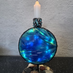 Op de afbeelding: Een handgemaakte kandelaar met een brandende witte kaars. De houder heeft een blauw en groen bolontwerp, versierd met kleine stenen en schelpen. De basis is cirkelvormig met een zanderige textuur.