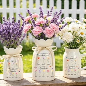 Könnte beinhalten: Drei cremefarbene Vasen gefüllt mit Blumen. Die linke Vase enthält Lavendel, die mittlere Vase rosa Nelken und die rechte Vase weiße Gänseblümchen. Jede Vase hat ein cremefarbenes Band und den Text "First Mom - Now Grandma".
