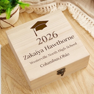 Pode incluir: Uma caixa de madeira clara com um gráfico de chapéu de formatura e o ano de 2026. A caixa é gravada com o nome Zakaiya Hawthorne, Westerville North High School e Columbus, Ohio. Possui um fecho de metal.