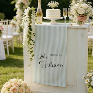 May include: A wedding bar decorated with a blue and white striped runner bearing the text "The Cheers to Williams." The bar is adorned with a cake, champagne, and flowers. A bouquet of white roses is visible in the background.